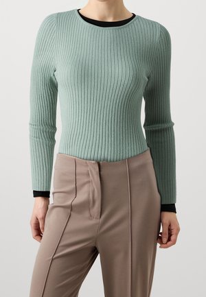 Vrouw draagt een aansluitend, geribbeld mintgroen shirt met lange mouwen over een zwart ondershirt, gecombineerd met beige, op hoge taille gesneden, op maat gemaakte broek.