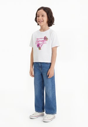 Kind mit lockigem Haar lächelt, trägt ein weißes T-Shirt mit Blumenmuster, blaue weite Jeans und weiße Turnschuhe, steht vor weißem Hintergrund.