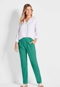 Witte blouse met lange mouwen en groene elastische taillebroek. De broek heeft zijzakken. Model draagt sandalen met straps.