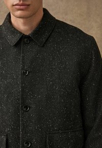 Palton negru cu model herringbone, realizat dintr-un amestec de lână texturat, cu închidere prin butoane și două buzunare laterale. Prezența unor mici puncte albe în întregul material.