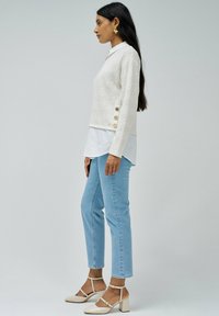 Pull crème texturé avec des accents de bouton sur le côté, superposé sur une chemise blanche ; associé à un jean bleu clair et des chaussures à talons carrés nude.