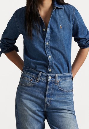 Kvinde iført langærmet, knappet blå denimskjorte og højtaljede blå jeans med hænderne bag ryggen.