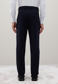 Man in een marineblauwe broek en zwarte schoenen, staande op een lichtgrijze vloerbedekking tegen een effen beige muur, gezien van achteren.