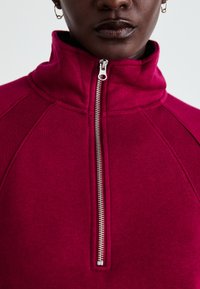 Pull en fuchsia avec col montant, fermeture éclair à l'avant et manches raglan. Texture lisse avec coutures visibles et quincaillerie de fermeture éclair en acier inoxydable.