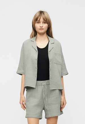 Jeune femme debout, portant une chemise boutonnée à manches courtes texturée gris clair et un short assorti à cordon sur un haut noir.