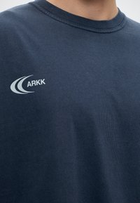 Άτομο που φοράει μπλούζα σε ναυτικό μπλε με λευκό λογότυπο ARKK στο αριστερό μέρος του στήθους.