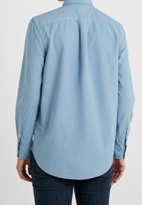 Camicia button-up azzurro chiaro, maniche lunghe, orlo curvo e colletto standard, realizzata in tessuto morbido con cuciture e bottoni ben visibili.