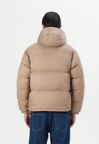 Puffjakke i lys beige med hætte, quiltet polstring og elastiske manchetter, der er parret med blå jeans, vist fra ryggen.
