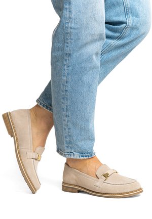 Beige Wildleder-Slipper mit gestepptem Design, goldfarbenen Beschlägen und einer profilierten Gummisohle, getragen zu hellblauen, umgekrempelten Jeans.
