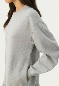 Grauer Kapuzenpullover mit Rundhalsausschnitt, langen Ärmel und Seitentaschen. Hergestellt aus weichem, strukturiertem Stoff mit sichtbaren Nähdetails.