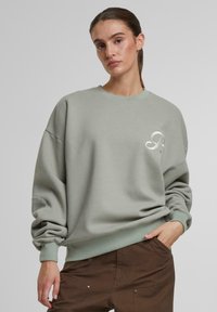 Lys grønn oversized sweatshirt i myk stoff med ribbet hals og mansjetter, med en subtil logo på brystområdet.