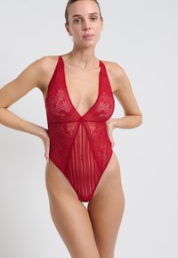 Body rosso in pizzo con scollatura a V profonda, motivi floreali intricati e pannelli a strisce trasparenti. Spalline regolabili e aperture delle gambe alte.