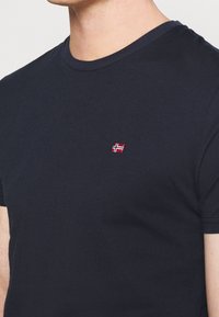 Marinblå bomullströja med rund hals, korta ärmar och en liten broderad logotyp i rött och vitt på vänster bröst.