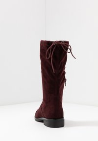 Botte haute en daim bordeaux avec un bout arrondi, un cordon élastique en haut et un talon bas noir. Dotée d'une fermeture éclair latérale pour plus de facilité.