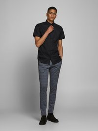 Chemise noire à manches courtes avec boutons, associée à un pantalon slim de couleur grise à carreaux et des bottines noires. Tissu lisse, coupe ajustée.
