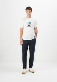 Hvid bomulds T-shirt med en tegneseriefigur med briller og ordet "GENIUS." Sammen med marineblå sweatpants og beige sneakers.
