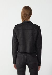 Zwarte nep-suède cropped jack met een kraag, lange mouwen en schouderdetails, gecombineerd met donkere jeans. Matte textuur.