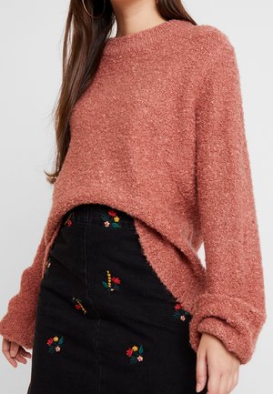 Femme portant un pull rose texturé à manches longues et col montant, associé à une jupe noire ornée de petites broderies florales colorées.