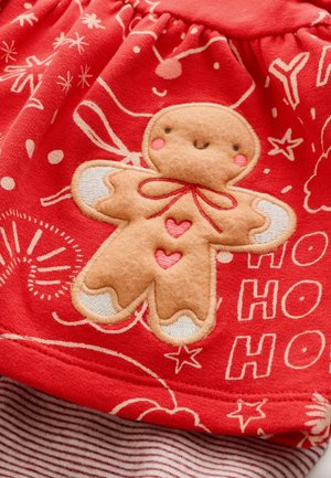 Röd tyg med en pepparkaksgubbe-applikation, med ljusbruna detaljer, röda accenter och vita höjdpunkter. Bakgrunden har festliga vita mönster.