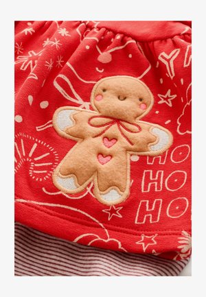 Tessuto rosso con un'applicazione a forma di uomo di pan di zenzero, caratterizzata da dettagli marrone chiaro, accenti rossi e evidenziature bianche. Lo sfondo ha motivi festivi bianchi.