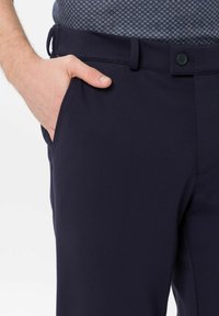 Homme portant un pantalon bleu marine avec une main dans la poche avant, bouton et passants de ceinture visibles, et une chemise à motifs rentrée.