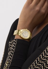 Guld-tone armbåndsur med en smykkebezel og skeleton urskive på et håndled iført en sort langærmet top med guldstuder.