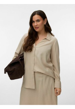 Ung kvinne med langt brunt hår iført en beige skjorte med knapper og skjørt, holding en stor brettet mørk brun veske i venstre hånd.