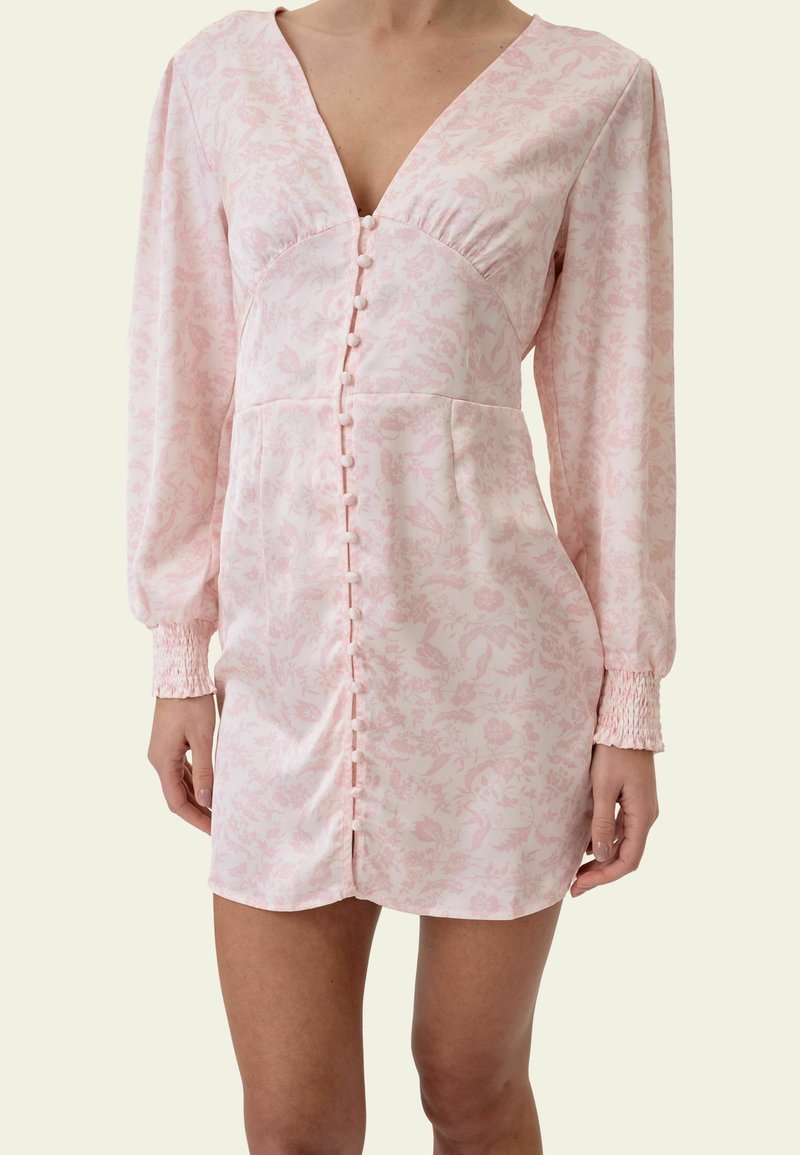 Femme portant une mini-robe rose pâle à manches longues avec un motif floral, une fermeture boutonnée devant et des poignets smockés.