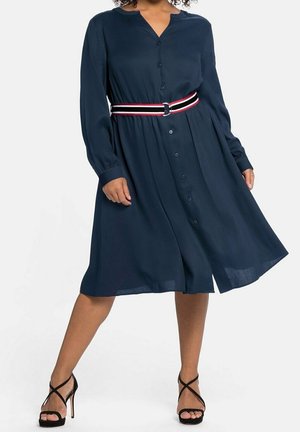 Femme portant une robe chemise bleue marine à manches longues avec une ceinture rayée et des sandales noires à talons hauts, debout, jambes écartées.