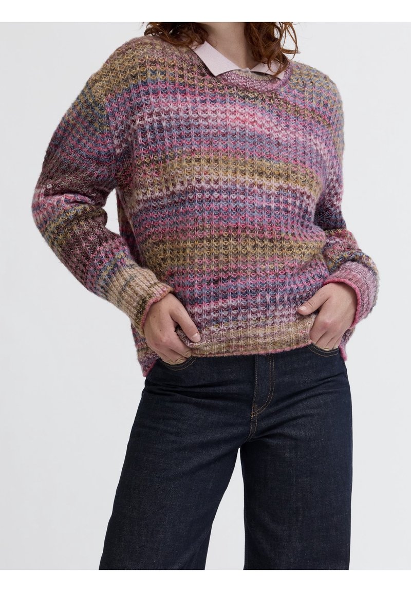 Personne portant un pull en maille multicolore avec des rayures roses, violettes et marron et un jean bleu foncé, les mains dans les poches.