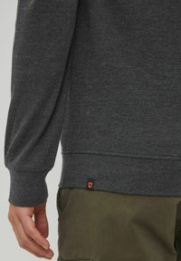 Charcoal gray sweatshirt with ribbed cuffs and hem, featuring a small logo tag in orange at the lower side, paired with olive green pants.