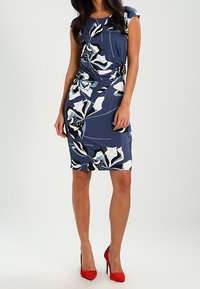 Robe fleurie à manches courtes volantées en bleu marine avec des motifs blancs et turquoise. Coupe ajustée avec fronces ; assortie à des talons hauts rouges. Tissu lisse et extensible.