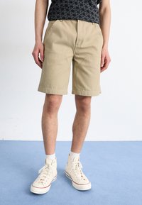 Mand iført beige knælange shorts, sort mønstrets skjorte, hvide sokker og cremefarvede høje sneakers, som står på blå gulv.