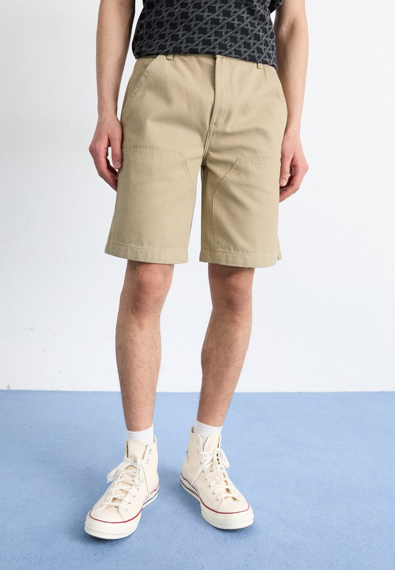 Mand iført beige knælange shorts, sort mønstrets skjorte, hvide sokker og cremefarvede høje sneakers, som står på blå gulv.
