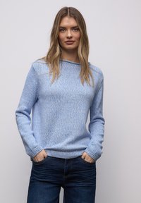 Femme aux cheveux longs et blonds porte un pull en maille bleu clair et un jean foncé, debout les mains dans les poches devant un fond gris uni.