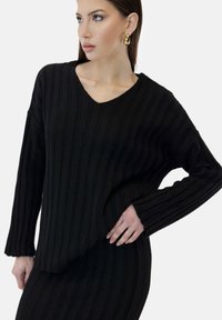 Sort strikket sweater med v-hals og lange ærmer med ribbet tekstur og lodrette striber. Stoffet ser blødt og tekstureret ud.