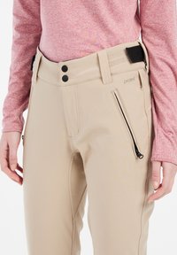 Beige Hosen mit einer eleganten Gestaltung, ausgestattet mit einer Reißverschlusstasche, zwei schwarzen Knöpfen und einem strukturierten Taillenbund. Der Stoff wirkt glatt und robust.