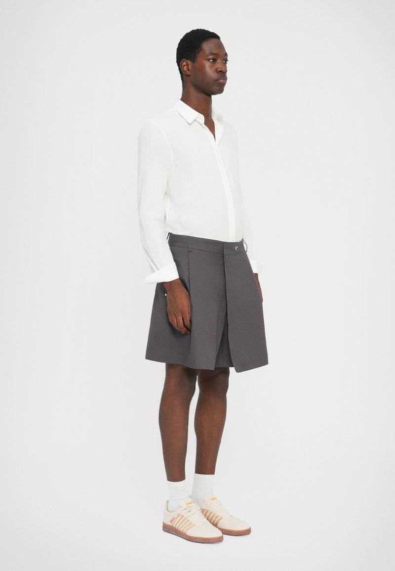 Grijze rok met plooien, wit overhemd met knopen, witte sokken en sneakers met beige accenten. Model staat tegen een effen witte achtergrond.