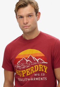 Rød bomulds T-shirt med et grafisk design af bjerge og en solnedgang i orange og gul med teksten "SUPERDRY MFG. CO QUALITY GARMENTS."