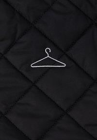 Tissu matelassé noir avec un motif en losange, présentant un design de cintre brodé blanc au centre. Texture lisse et couture uniforme.