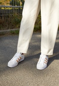 Vita sneakers med tre persikofärgade ränder, gummitåhätta och snörning. Bärs med löst sittande krämfärgade byxor och står på en strukturerad asfalt yta.