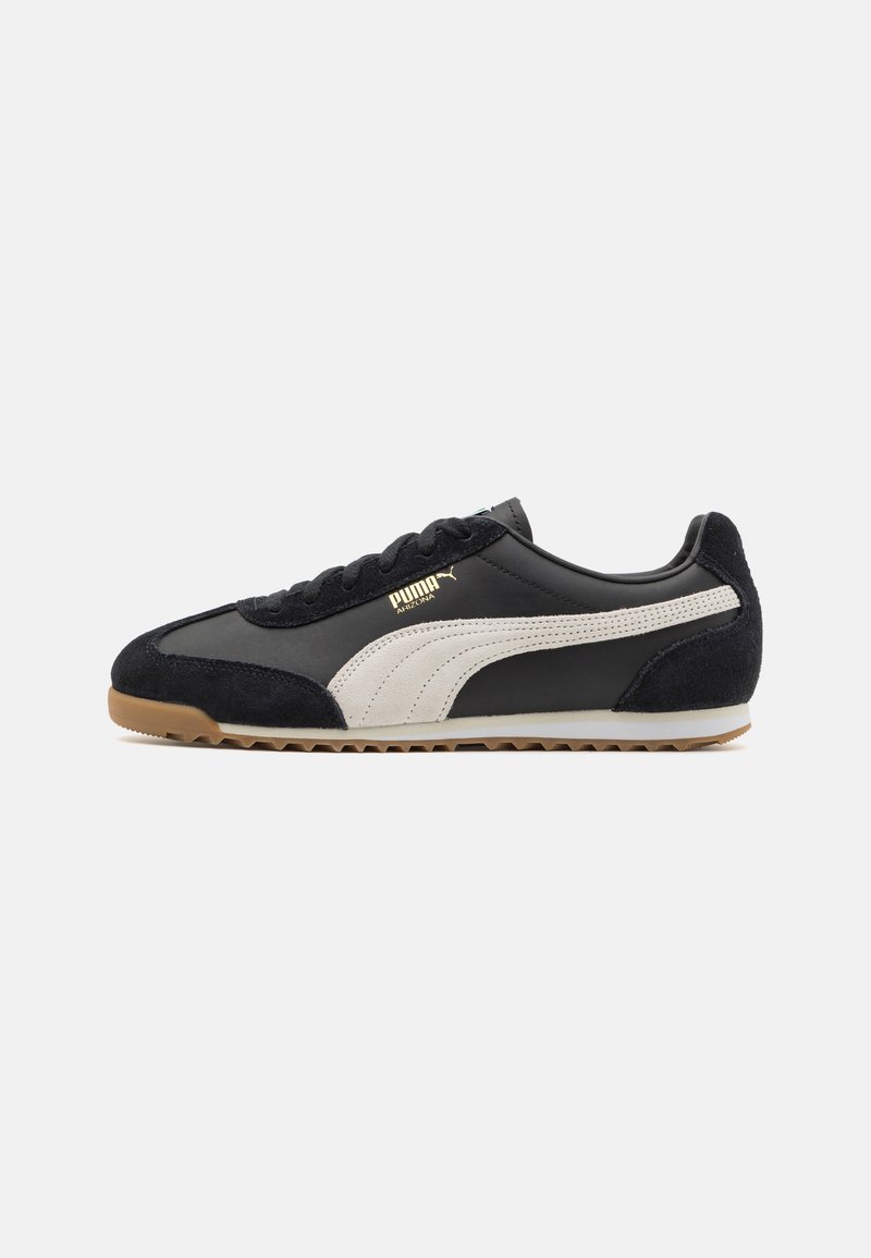 Adidașii Puma în nuanțe de negru și alb, realizați din material din suede și piele, cu un design de profil scăzut, accente texturate și talpă din cauciuc de tip gumă. Detaliu cu logo de culoare aurie.