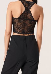 Top corto de encaje negro con borde en ondas y diseño de cuello halter, combinado con pantalones negros de talle alto. Se aprecia la textura detallada del encaje.