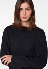 Pull en tricot noir avec une texture lisse, un col large et des manches longues. Présente un motif en chevrons subtil et une coupe décontractée.