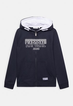 Granatowa bluza z kapturem na zamek, z białym podszyciem kaptura. Posiada przednie kieszenie oraz nadruk graficzny z napisem "KAPORAL JEANS" na piersi. Miękka faktura tkaniny.