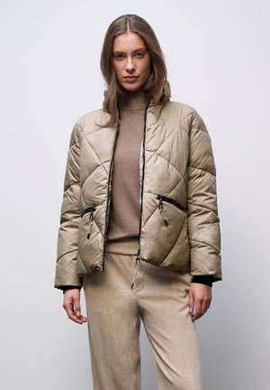 Giacca trapuntata beige con colletto alto, tasche con zip e dettagli neri, indossata sopra un maglione marrone a coste e pantaloni leggeri beige.