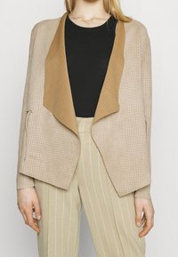 Veste beige perforée avec un col marron clair, ouverture frontale et manches longues, associée à un pantalon beige rayé. Design léger et texturé.