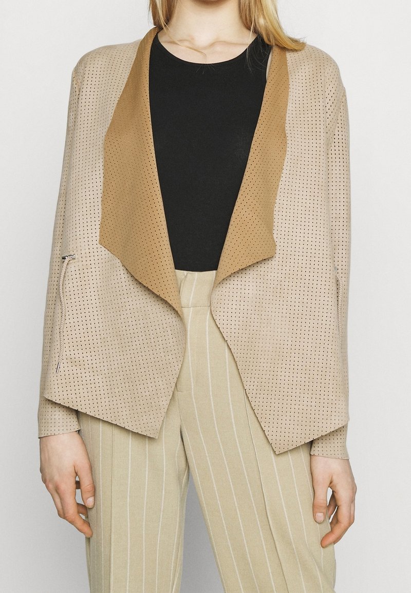 Veste beige perforée avec un col marron clair, ouverture frontale et manches longues, associée à un pantalon beige rayé. Design léger et texturé.