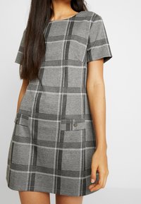 Grijs geruit jurkje met korte mouwen, een ronde halslijn, twee voorzakken met metalen accenten en een zachte textuur.