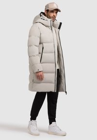 Lys grå puffjakke med hætte, sorte lynlåse og en lang silhuet. Båret over sorte bukser og hvide sneakers, med en beige kasket.